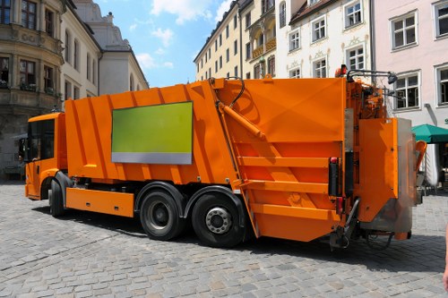 A large, modern orange refuse collection vehicle parked on a cobbled urban street with historic multi-storey buildings in the background. The vehicle features a smooth, metallic finish with a reflective yellow-green warning panel on its side. It has multiple compartments and hydraulic systems visible along the rear section, used for collecting and compacting rubbish. The scene is well-lit with natural daylight, highlighting the vehicle's bright colour and clean exterior. The setting suggests a busy town centre environment, typical of waste collection operations across Uxbridge, with paved pedestrian areas and nearby storefronts. Commercial Waste Removal Uxbridge operates vehicles like this for efficient rubbish removal services within the local area, supporting urban cleanliness and waste management needs in the town and surrounding postcode districts. The vehicle is designed for large capacity waste collection, with sturdy black tires, side mirrors, and functional loading apparatus clearly visible in the image, emphasizing professional rubbish removal capabilities in a typical busy street scene. The overall environment and vehicle details are captured clearly, aligning with commercial waste collection services offered by companies serving Uxbridge and its surrounding communities.