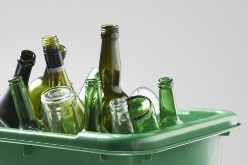 A close-up view of a green plastic recycling bin filled with various empty glass bottles of different shapes, sizes, and colours, predominantly green and brown. The bottles are arranged with some standing upright and others leaning, with their open mouths visible. The background is plain and light grey, highlighting the bottles and bin. The scene suggests a waste collection environment in an urban area, which could be associated with rubbish removal services such as those offered by Commercial Waste Removal Uxbridge in the local postcode area. The focus is on the glass bottles ready for recycling or disposal, relevant to waste management activity in a commercial or residential context in Uxbridge.
