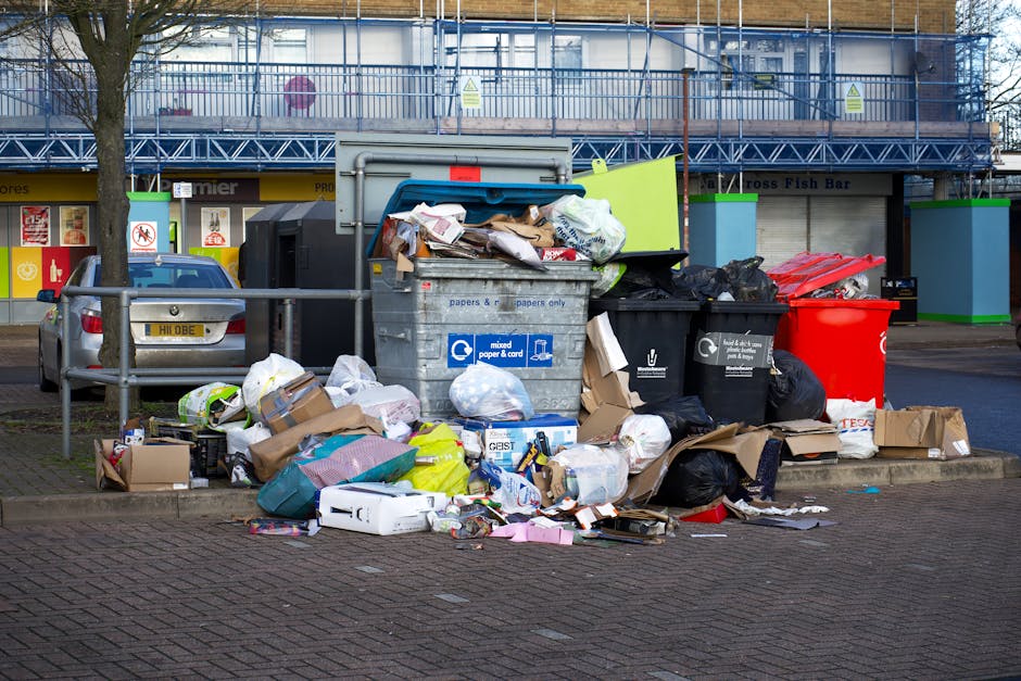 A pile of various discarded cardboard boxes, some flattened and others partially intact, rests against a rough, beige brick wall outdoors. The boxes are of different sizes, with visible printed labels